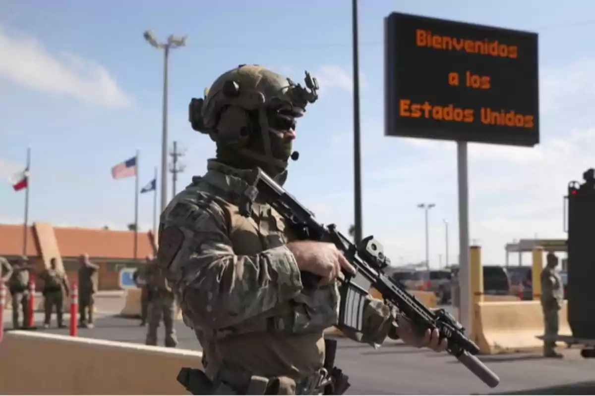 Un soldado armado con uniforme militar está de pie en un puesto fronterizo con un cartel de bienvenida a los Estados Unidos en el fondo.