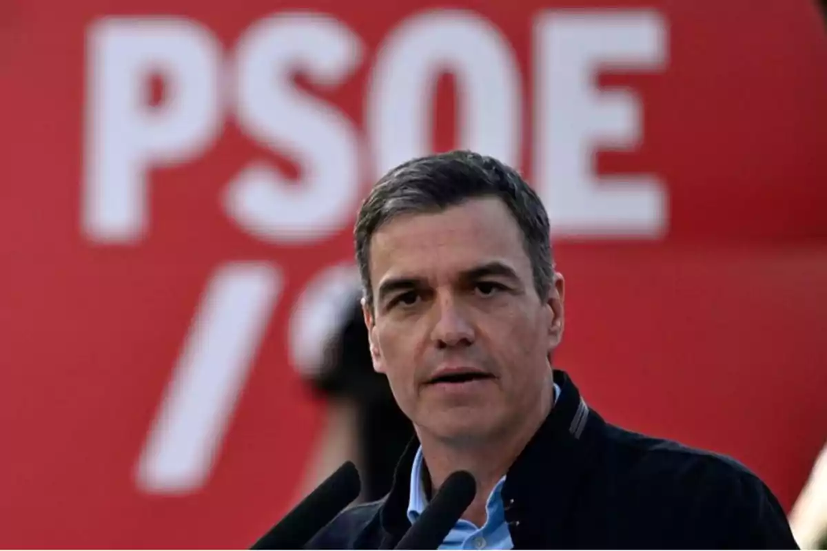 Un hombre hablando frente a un micrófono con un cartel rojo de fondo que tiene las letras 