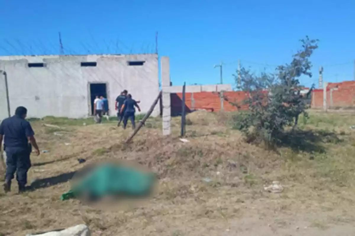 Un grupo de personas, incluyendo policías, se encuentra en un terreno con una construcción de fondo y un área de césped en primer plano.