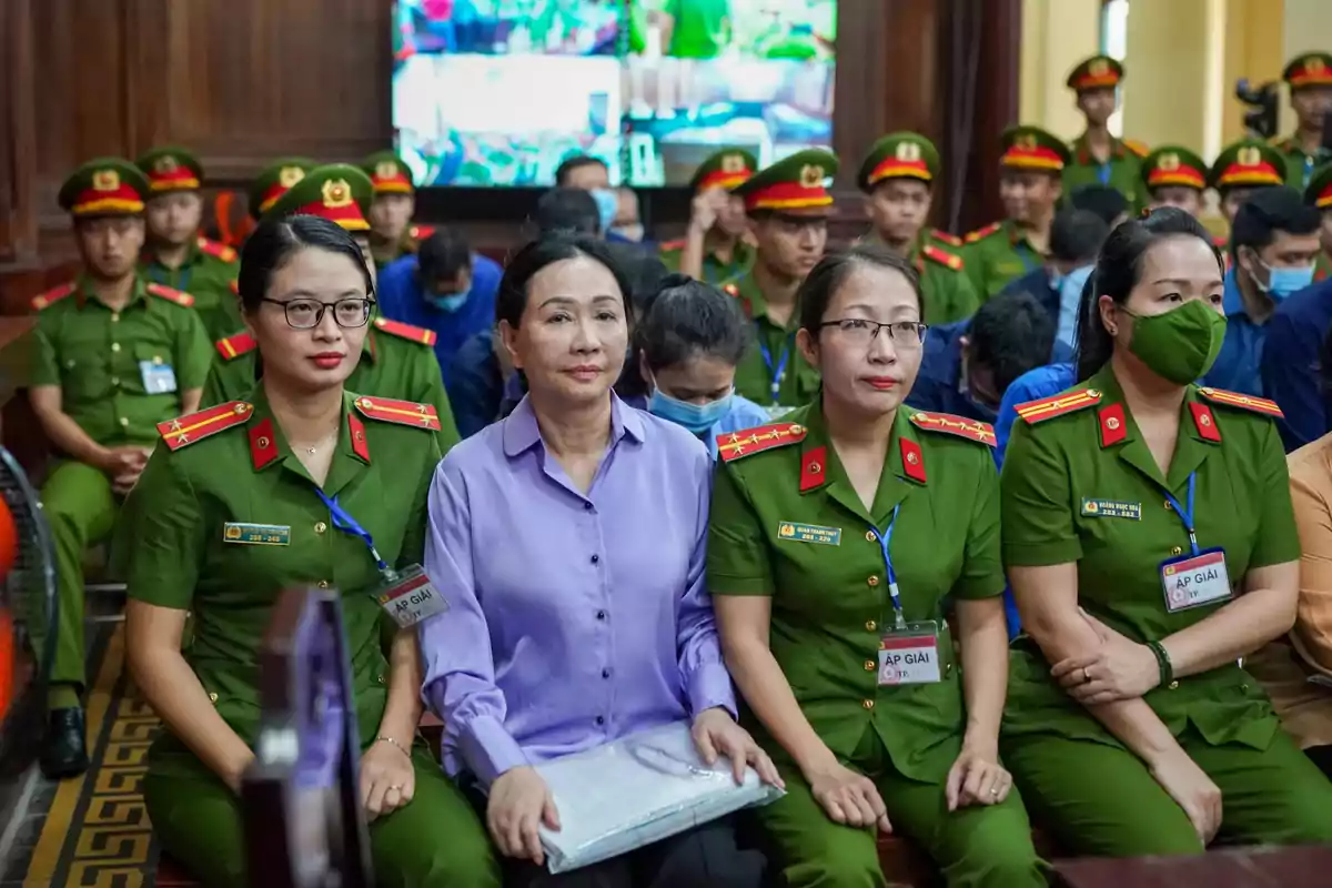 Un grupo de personas en uniforme verde y una mujer de camisa morada están sentadas en una sala con más personas al fondo.