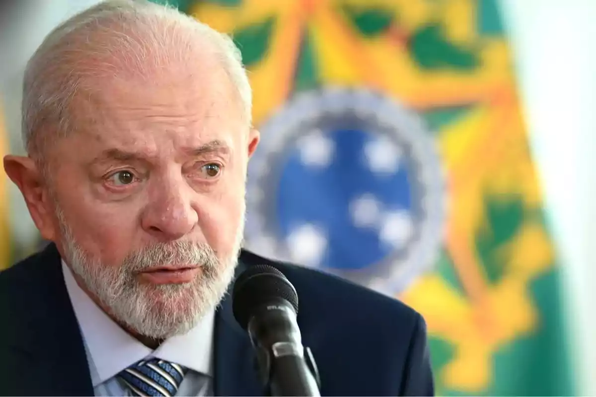 Un hombre mayor con barba blanca hablando frente a un micrófono con un fondo desenfocado de colores verde y amarillo.