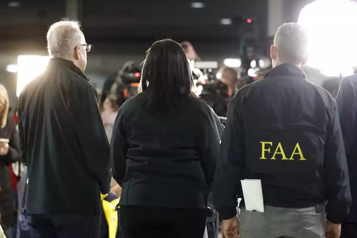 Personas de espaldas en un evento, una de ellas lleva una chaqueta con las letras FAA, rodeadas de cámaras y luces brillantes.