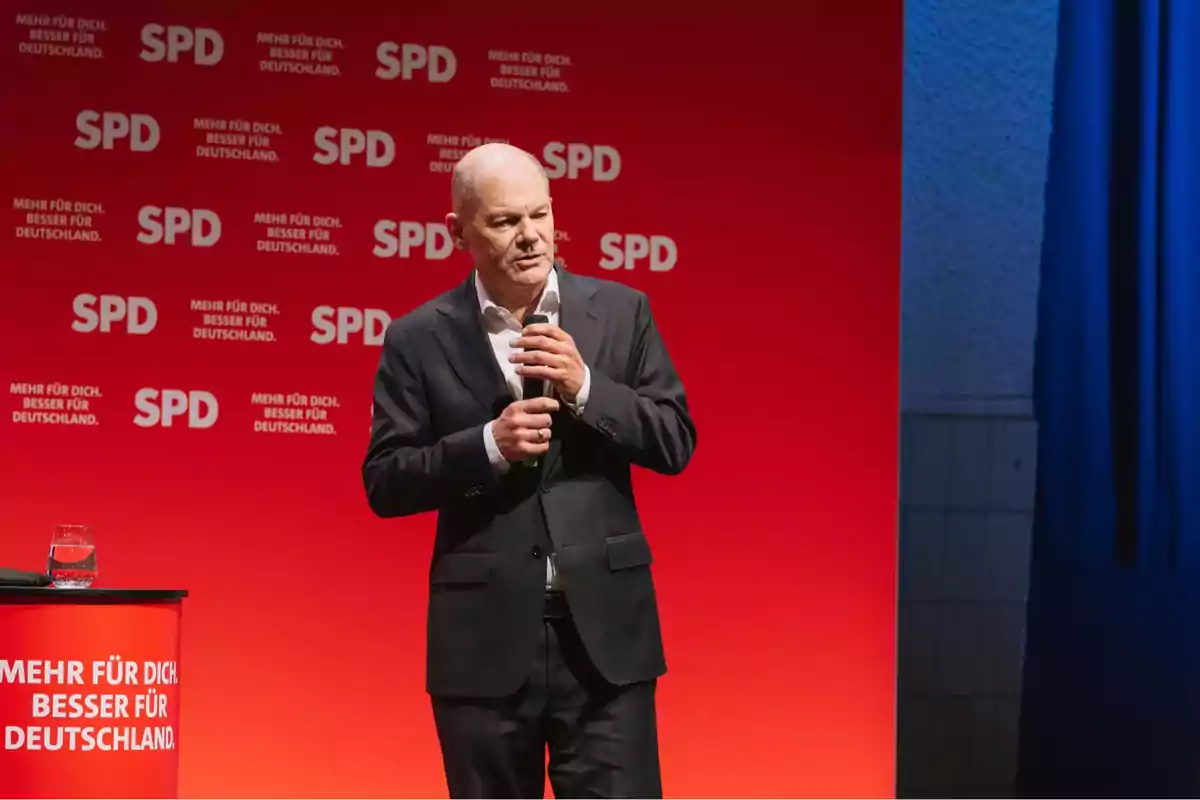 Un hombre en traje habla en un escenario con fondo rojo que tiene el logo del SPD y el lema 