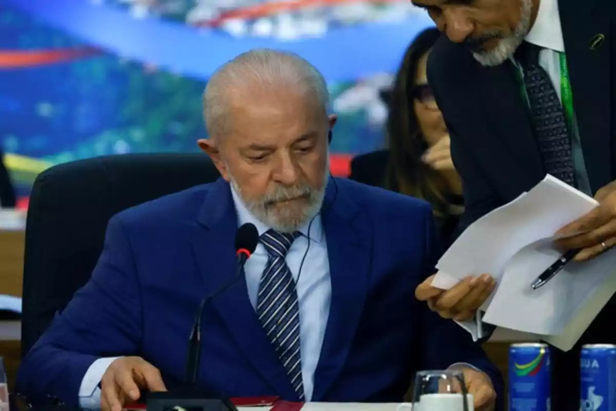 Un hombre de traje azul está sentado en una mesa con un micrófono frente a él mientras otra persona le muestra documentos.