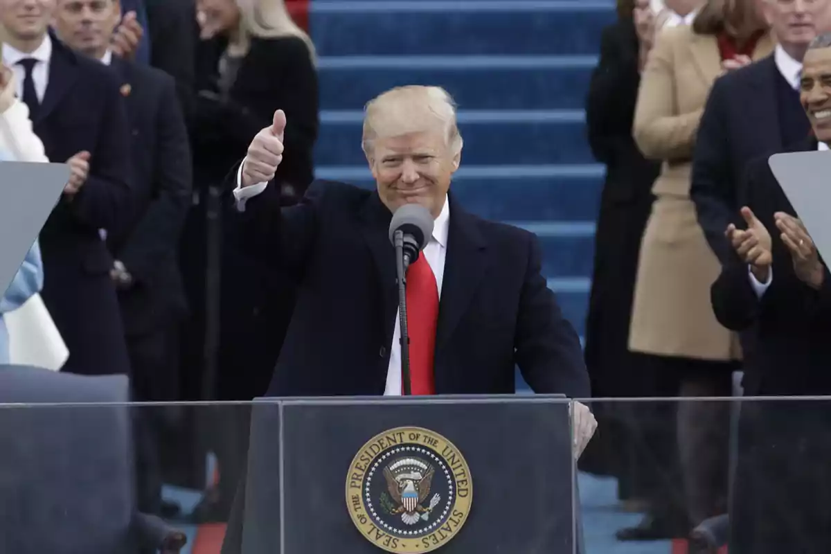 Un hombre con traje oscuro y corbata roja levanta el pulgar mientras está de pie frente a un podio con el sello presidencial de los Estados Unidos, rodeado de personas que aplauden.