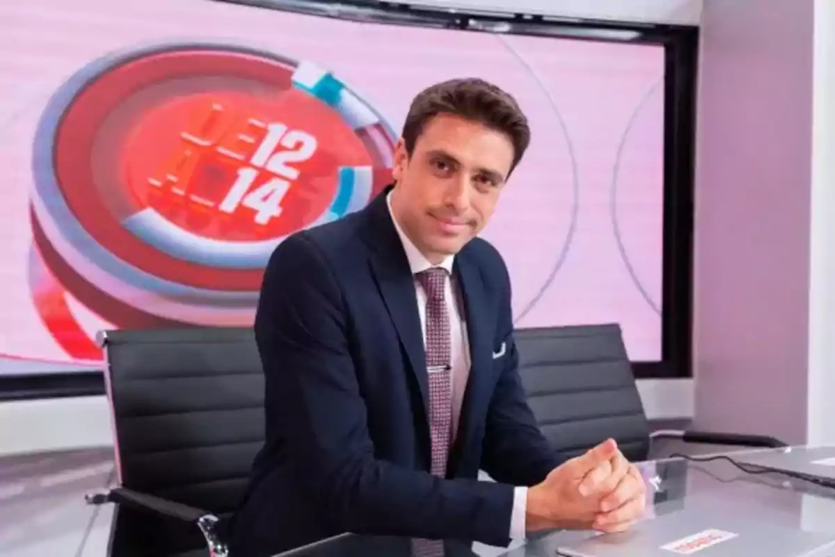 Un presentador de noticias sentado en un estudio de televisión con un fondo que muestra el logo de un programa.