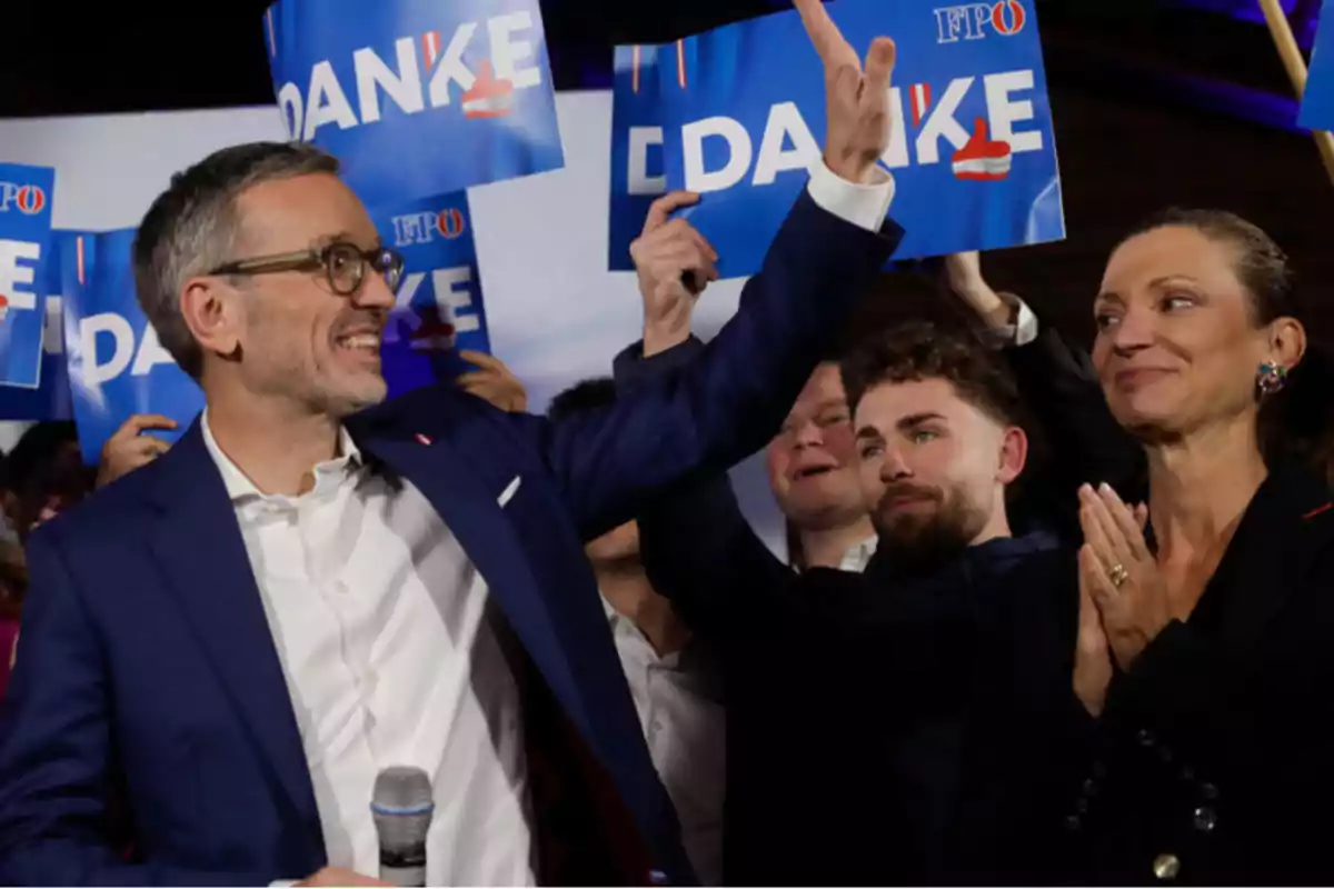 Un hombre con gafas y traje azul sonríe y saluda mientras sostiene un micrófono, rodeado de personas que sostienen carteles azules con la palabra "DANKE".