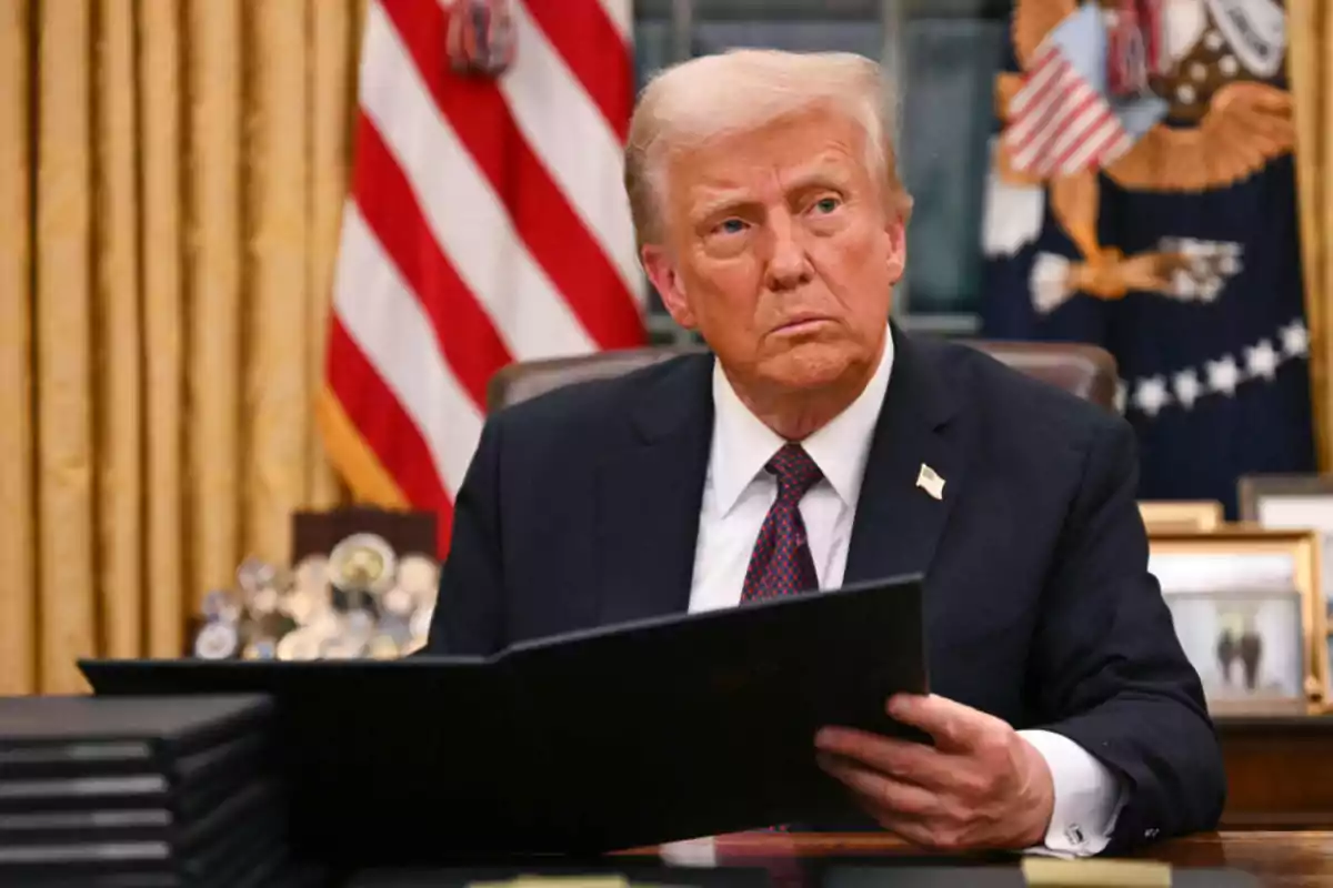 A man in a suit holding a folder in an office with flags and golden curtains in the background.