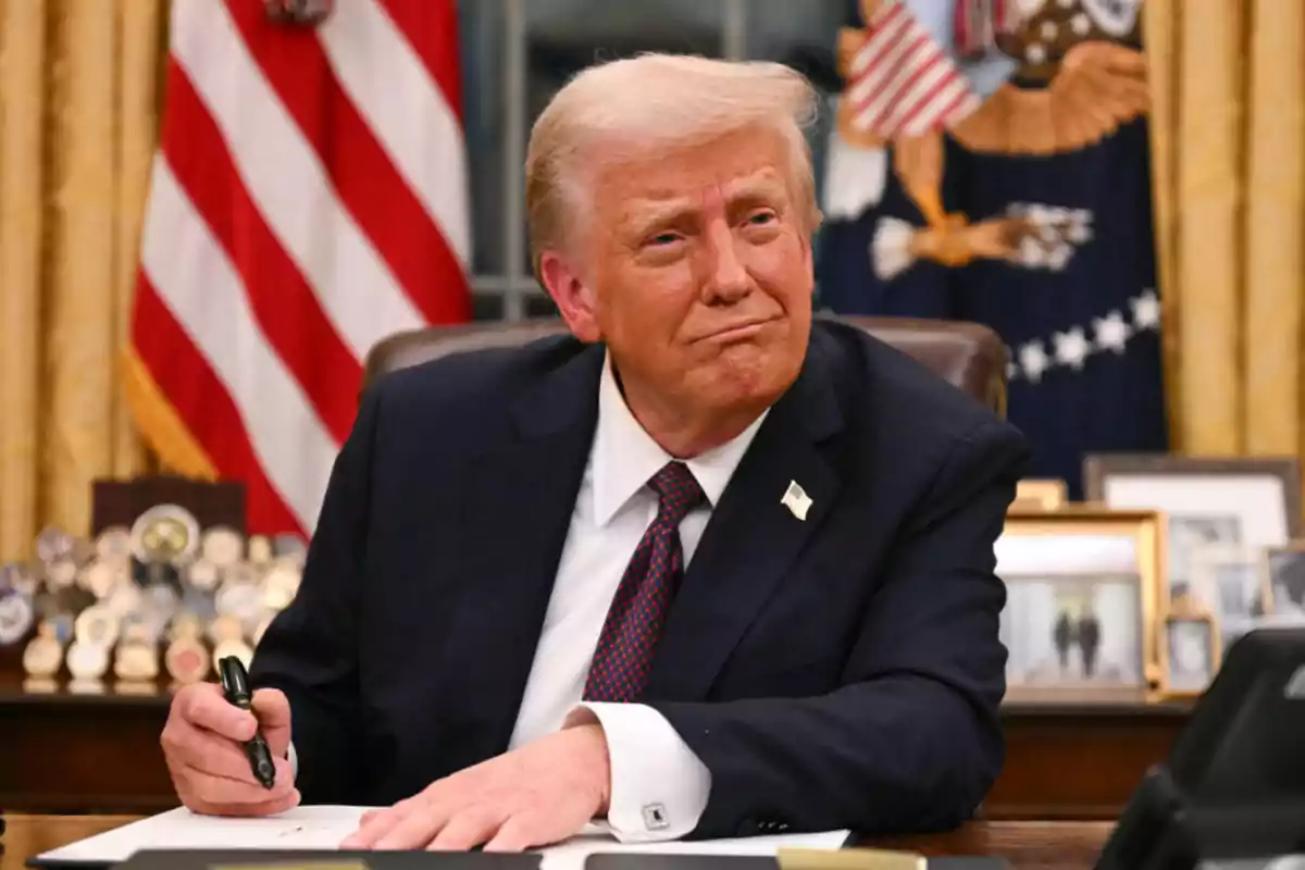 A man in a dark suit and red tie is sitting at a desk with an American flag in the background.