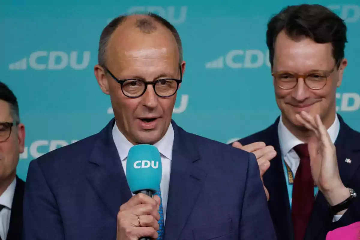 A man with glasses and a suit speaks into a blue microphone with the CDU logo while another person applauds in the background.