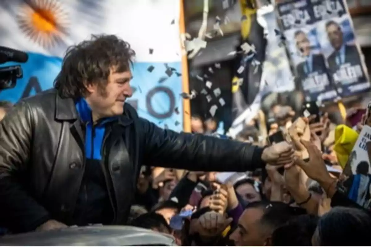 Un hombre con chaqueta de cuero saluda a una multitud que lo rodea mientras confeti cae a su alrededor y se ven banderas y carteles en el fondo.