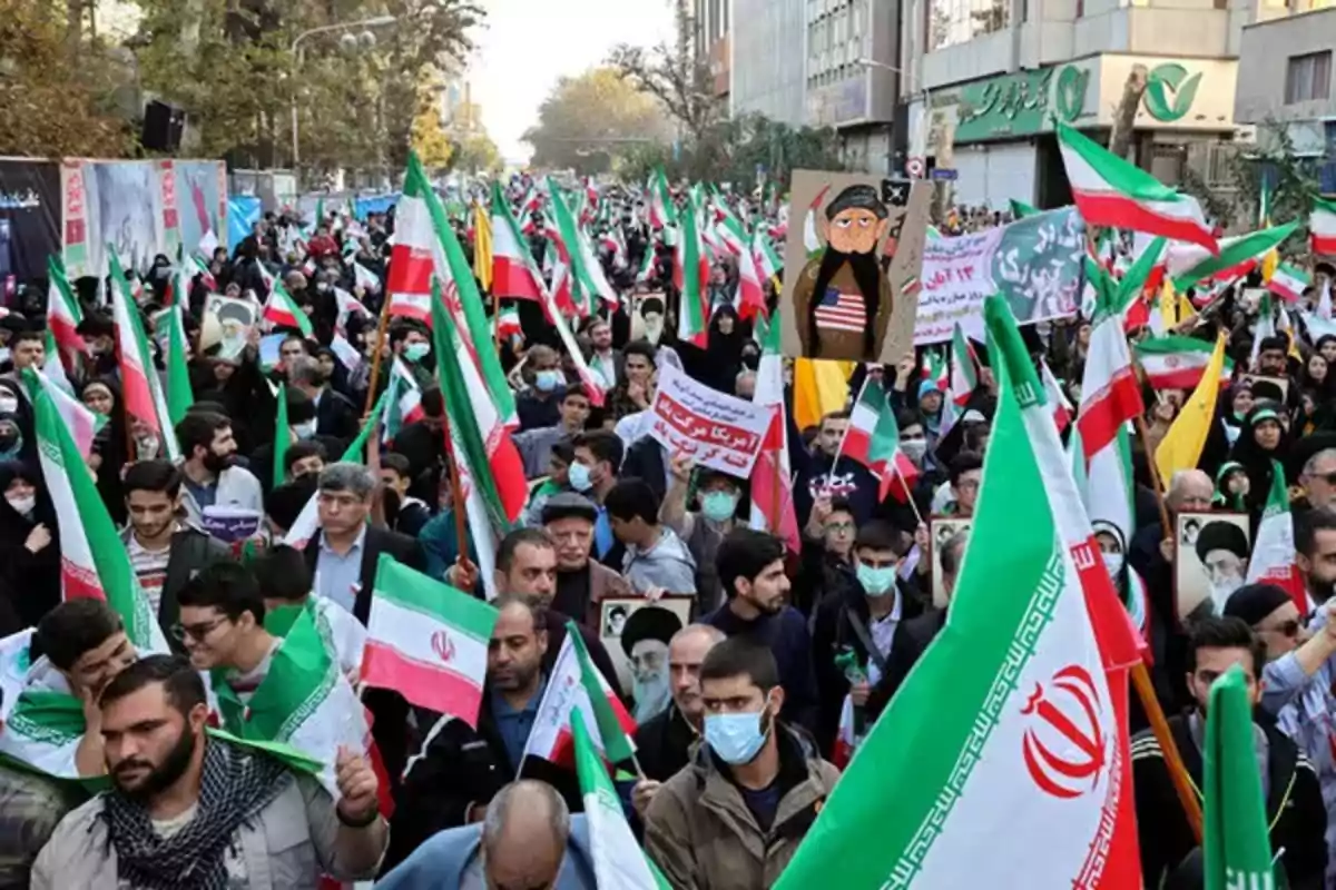 Una multitud de personas participa en una manifestación en la calle, portando banderas de Irán y carteles, algunos de ellos llevan mascarillas.