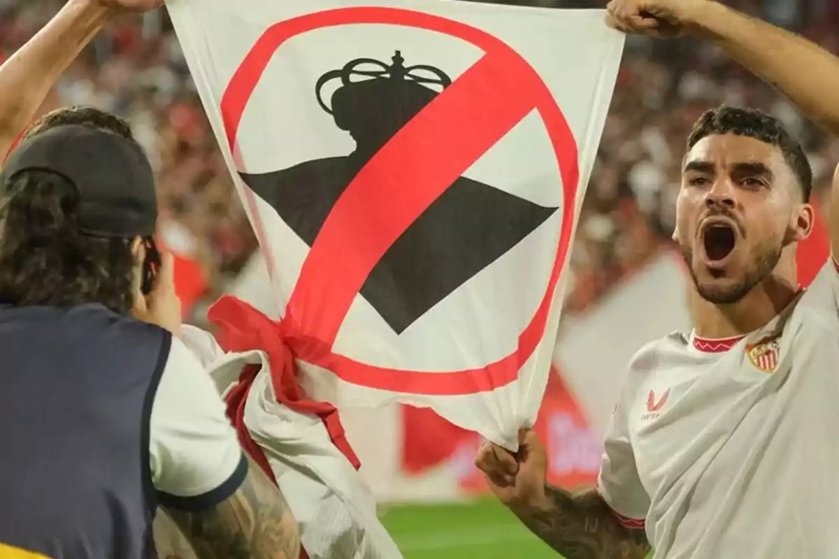 Un jugador de fútbol sostiene una bandera con un símbolo de prohibición sobre una corona en un estadio lleno de espectadores.