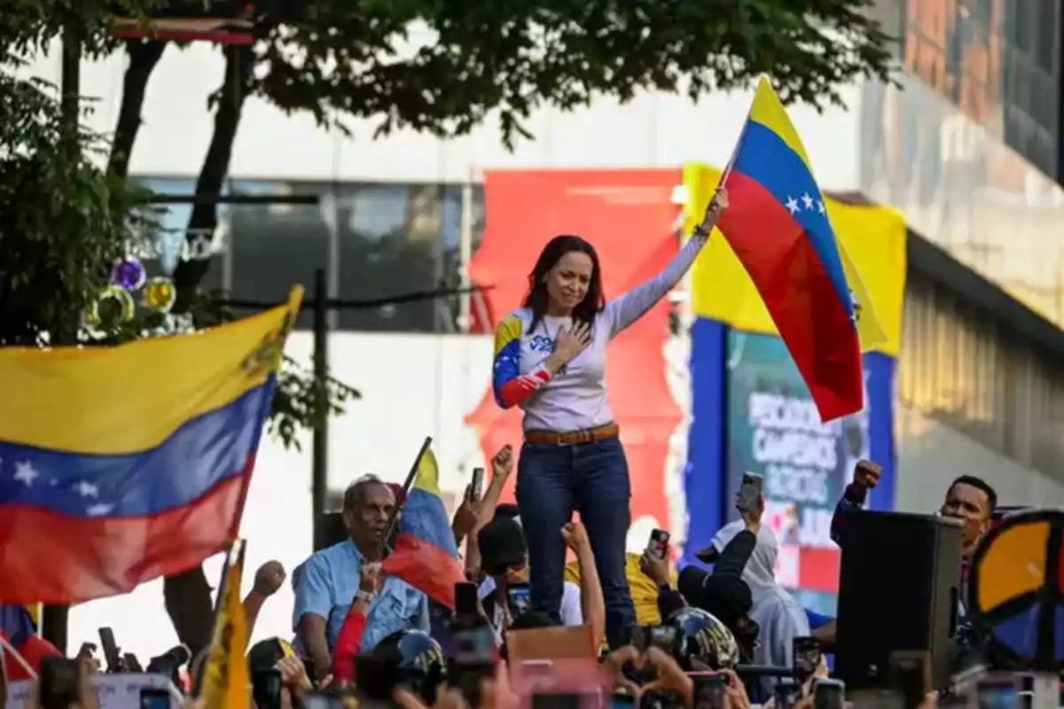 Una mujer sostiene una bandera de Venezuela mientras se encuentra de pie en un vehículo rodeada de una multitud que también lleva banderas venezolanas.