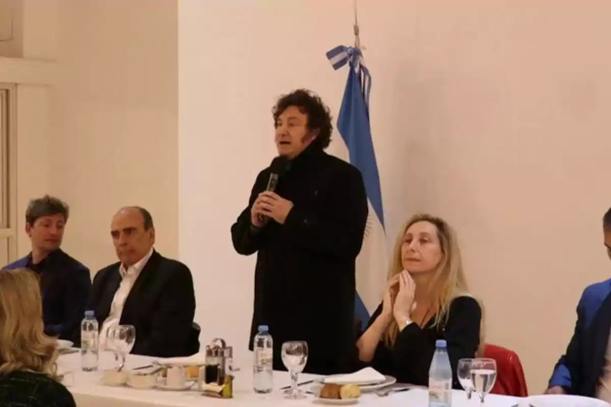 Una persona de pie hablando con un micrófono en una mesa con varias personas sentadas y una bandera en el fondo.