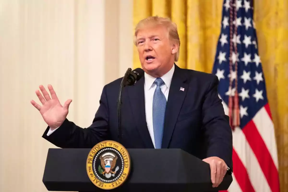 Un hombre hablando en un podio con un micrófono y una bandera de Estados Unidos al fondo.