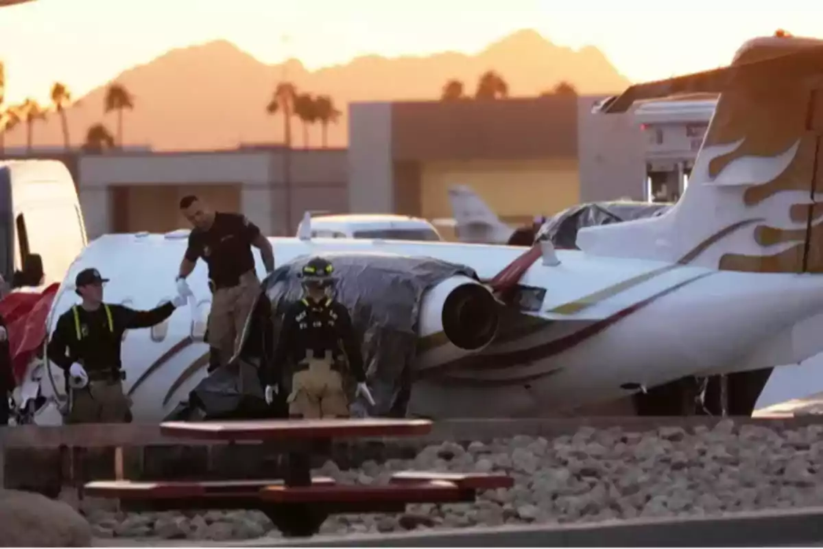 Un grupo de personas inspecciona un avión pequeño en un aeropuerto al atardecer con montañas y palmeras al fondo.