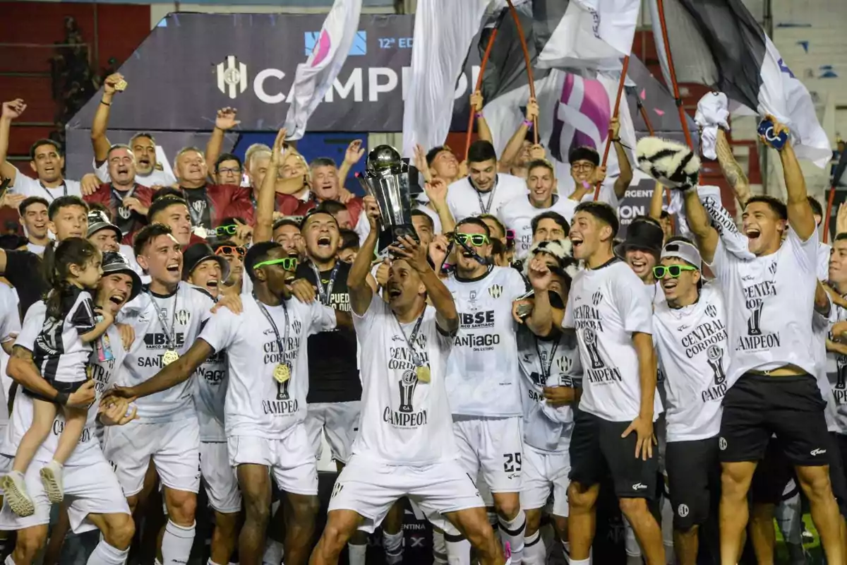 Un equipo de fútbol celebrando con un trofeo rodeado de aficionados y banderas.