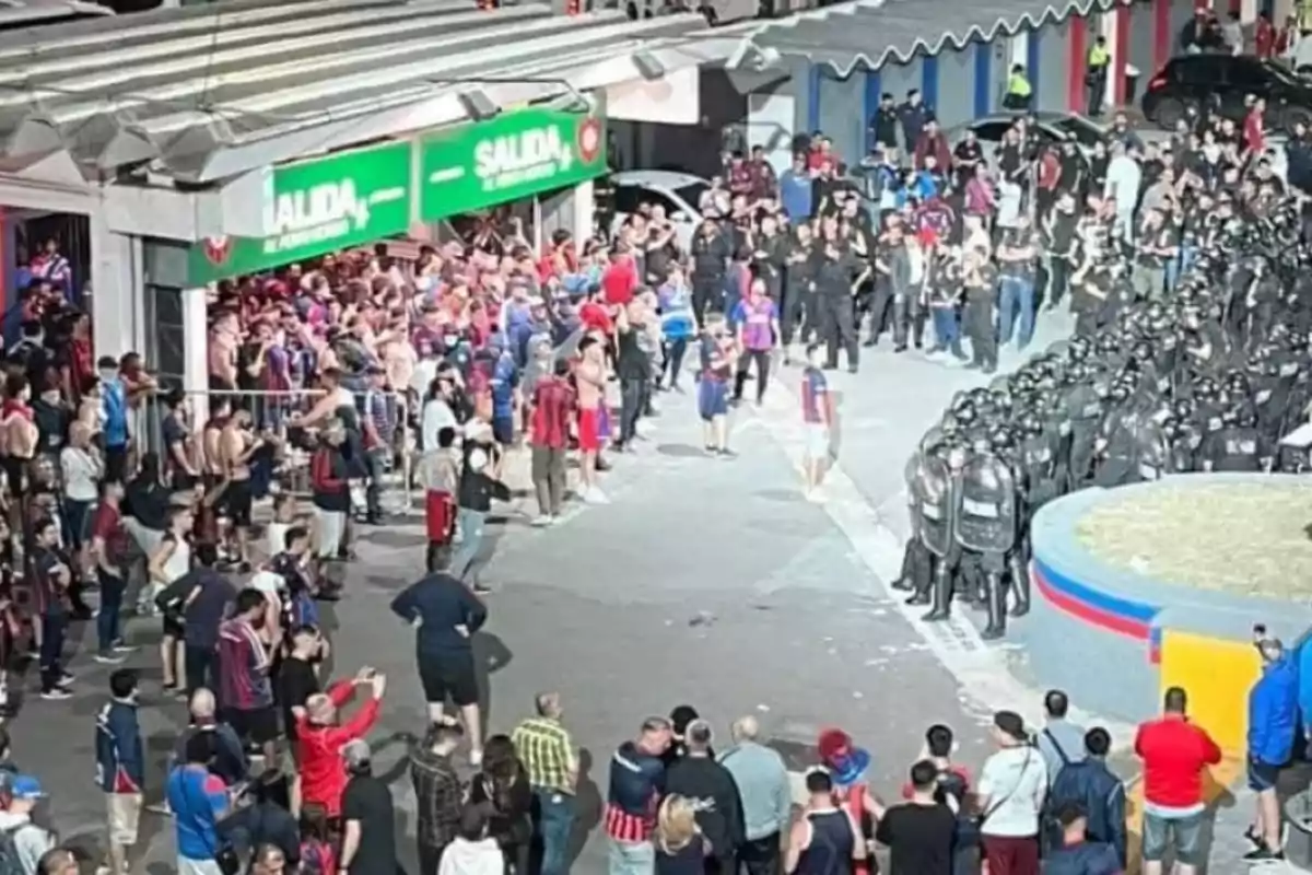 Un grupo de personas con camisetas de fútbol se encuentra frente a una salida de un estadio mientras un grupo de policías con equipo antidisturbios está alineado cerca.