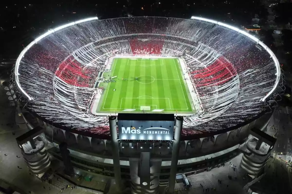 Vista aérea de un estadio de fútbol iluminado durante la noche con gradas llenas de espectadores y decoraciones en rojo y blanco.