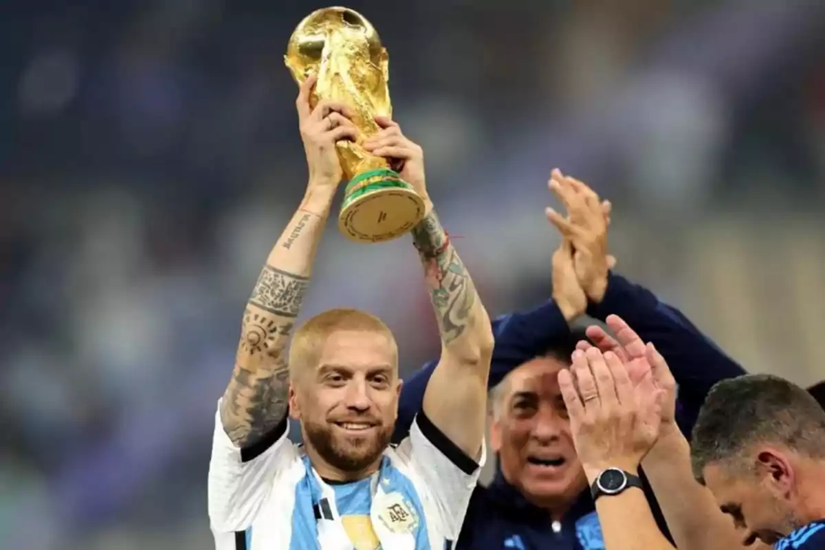 Un jugador de fútbol con uniforme de Argentina levanta un trofeo dorado mientras otras personas aplauden a su alrededor.