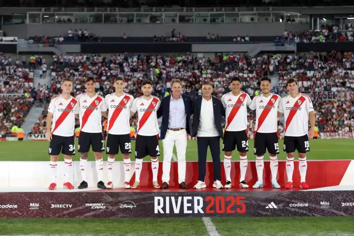 Un grupo de jugadores de fútbol y dos personas más posan en un estadio con una pancarta que dice "River 2025" al frente.