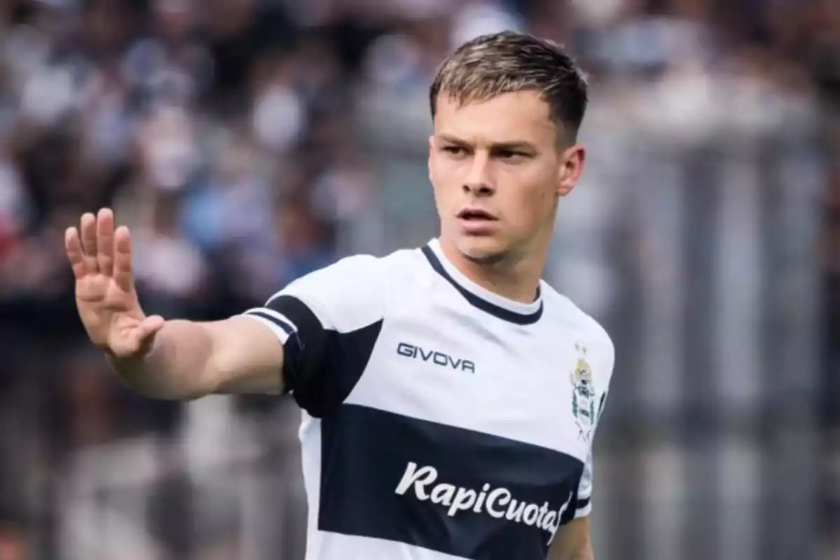 Un jugador de fútbol con uniforme blanco y negro levanta la mano en un gesto durante un partido.