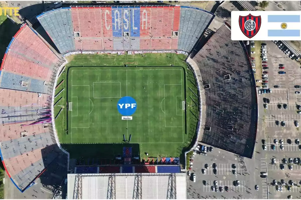 Vista aérea de un estadio de fútbol con gradas en colores rojo y azul, el campo de juego tiene un logo de YPF en el centro, hay un estacionamiento lleno de autos alrededor del estadio y en la esquina superior derecha se ven un escudo y una bandera.