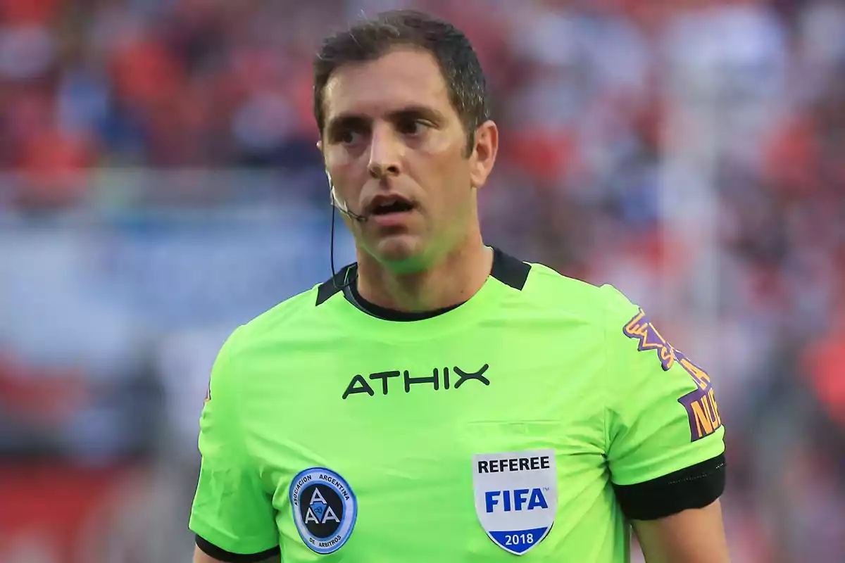 Un árbitro de fútbol con uniforme verde y escudo de la FIFA en el campo de juego.