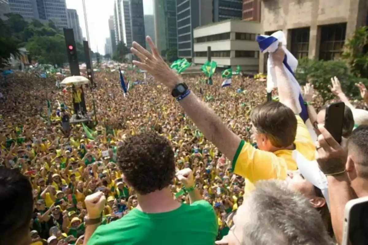 Una multitud de personas vestidas de amarillo y verde se reúne en una calle de una ciudad, ondeando banderas y levantando los brazos mientras un hombre en primer plano saluda desde una plataforma elevada.