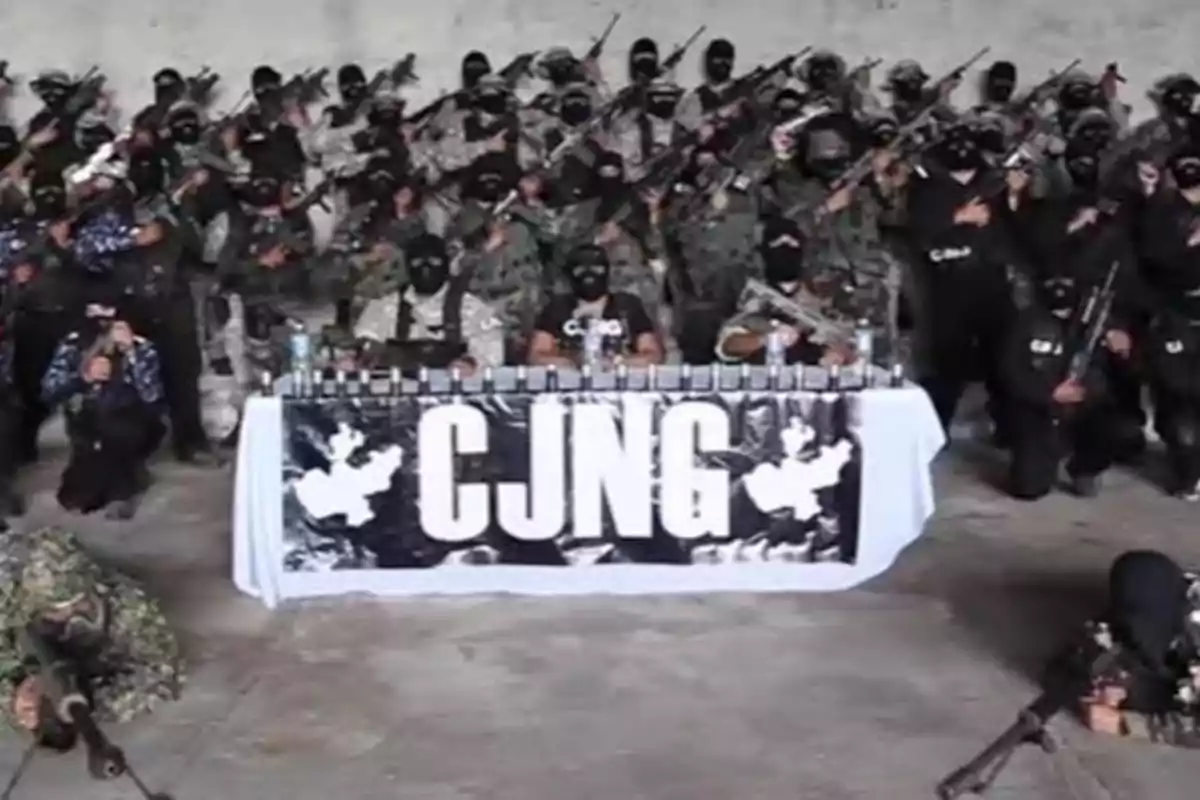 Un grupo de personas enmascaradas vestidas de camuflaje y negro posan con armas detrás de una mesa con un cartel que tiene las letras "CJNG".