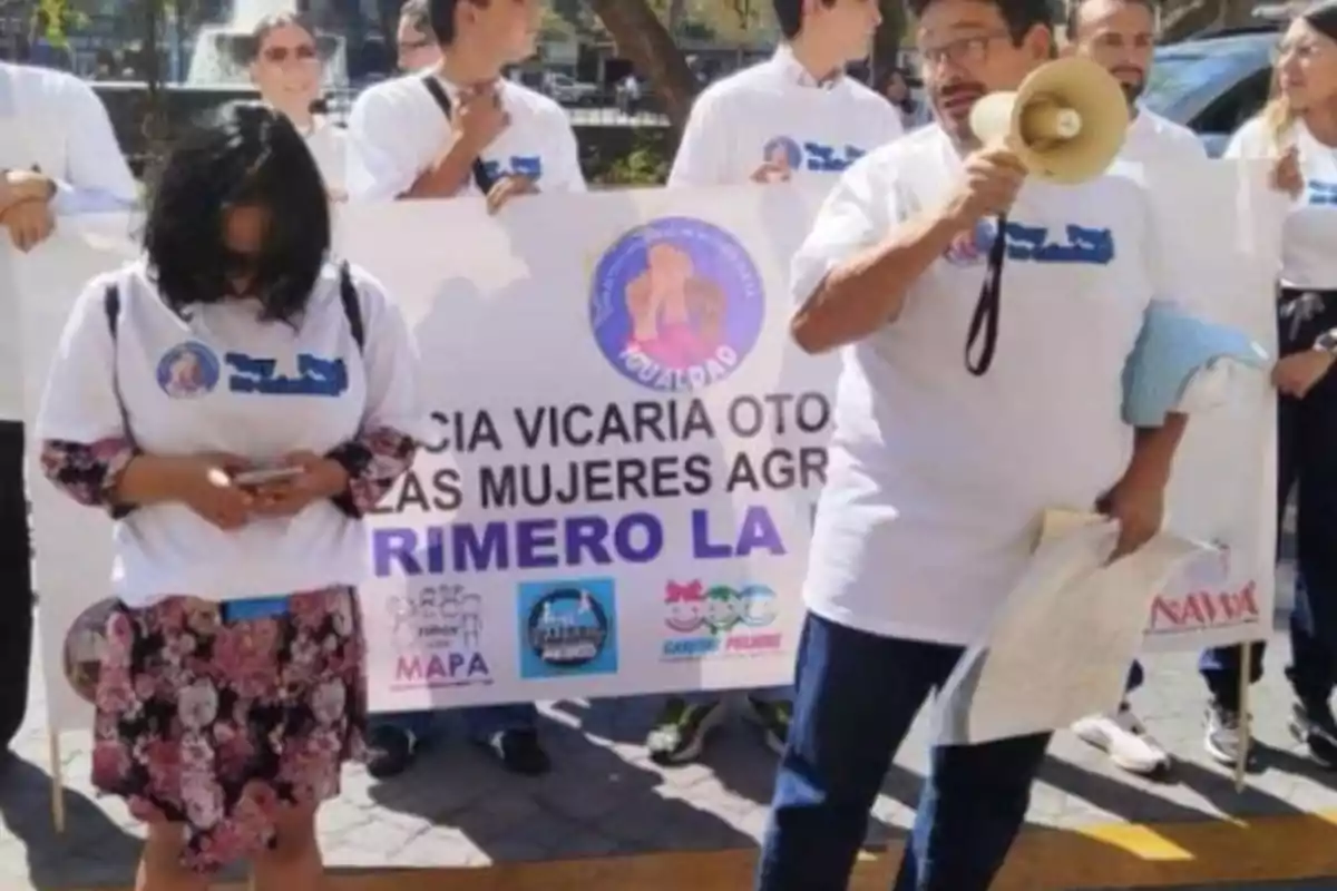 Un grupo de personas con camisetas blancas sostiene una pancarta durante una manifestación; una persona usa un megáfono.