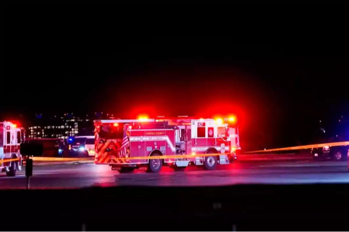 Un camión de bomberos con luces rojas encendidas está estacionado en una calle por la noche, rodeado de cinta de seguridad amarilla y otros vehículos de emergencia.