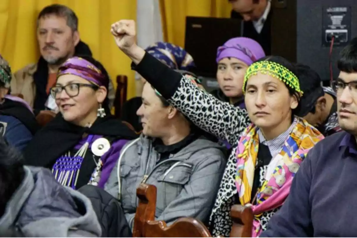 Un grupo de personas, algunas con pañuelos en la cabeza, se encuentran sentadas en una sala; una de ellas levanta el puño en señal de protesta.