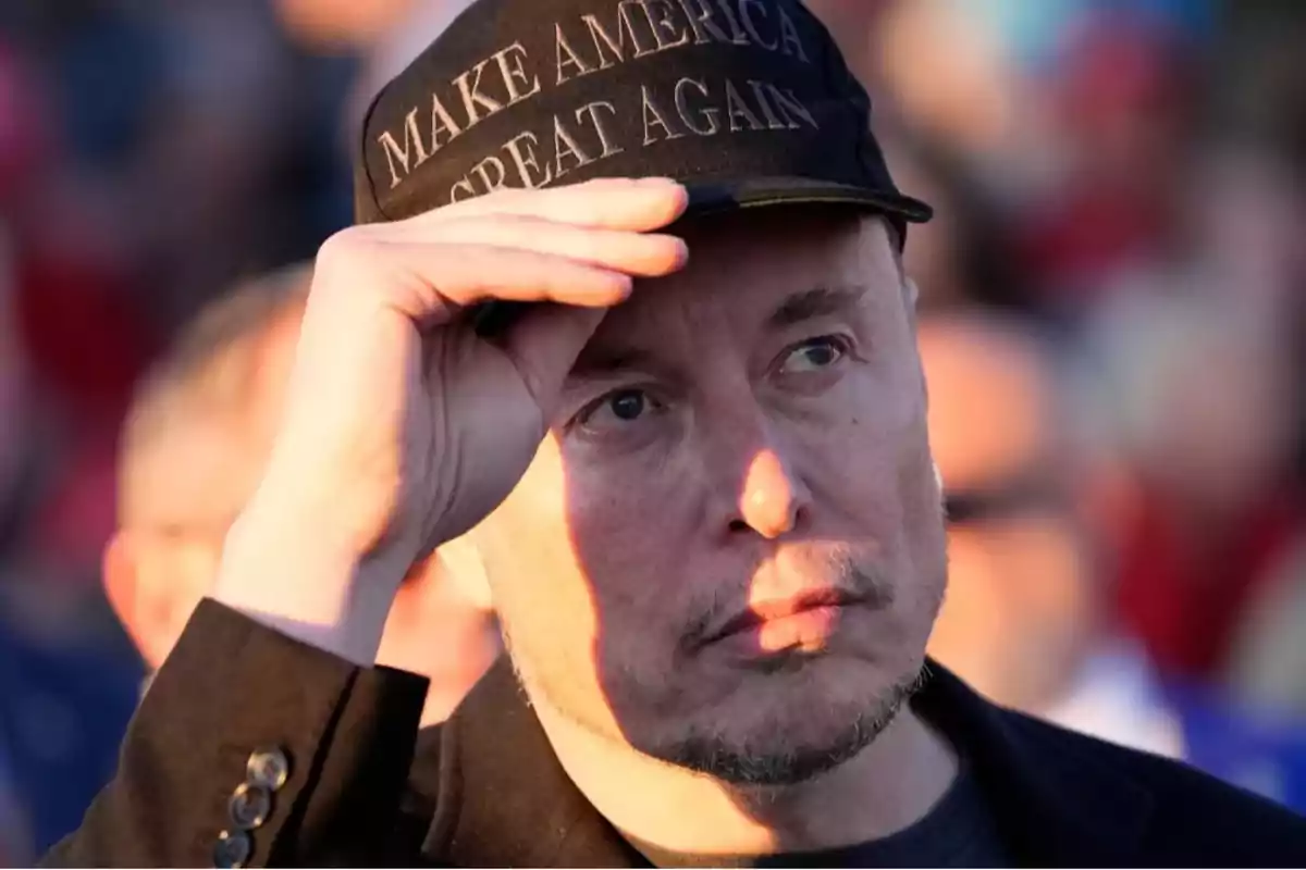 Un hombre con gorra negra que tiene un mensaje en letras blancas, levantando la mano para ajustar la visera, con fondo desenfocado.