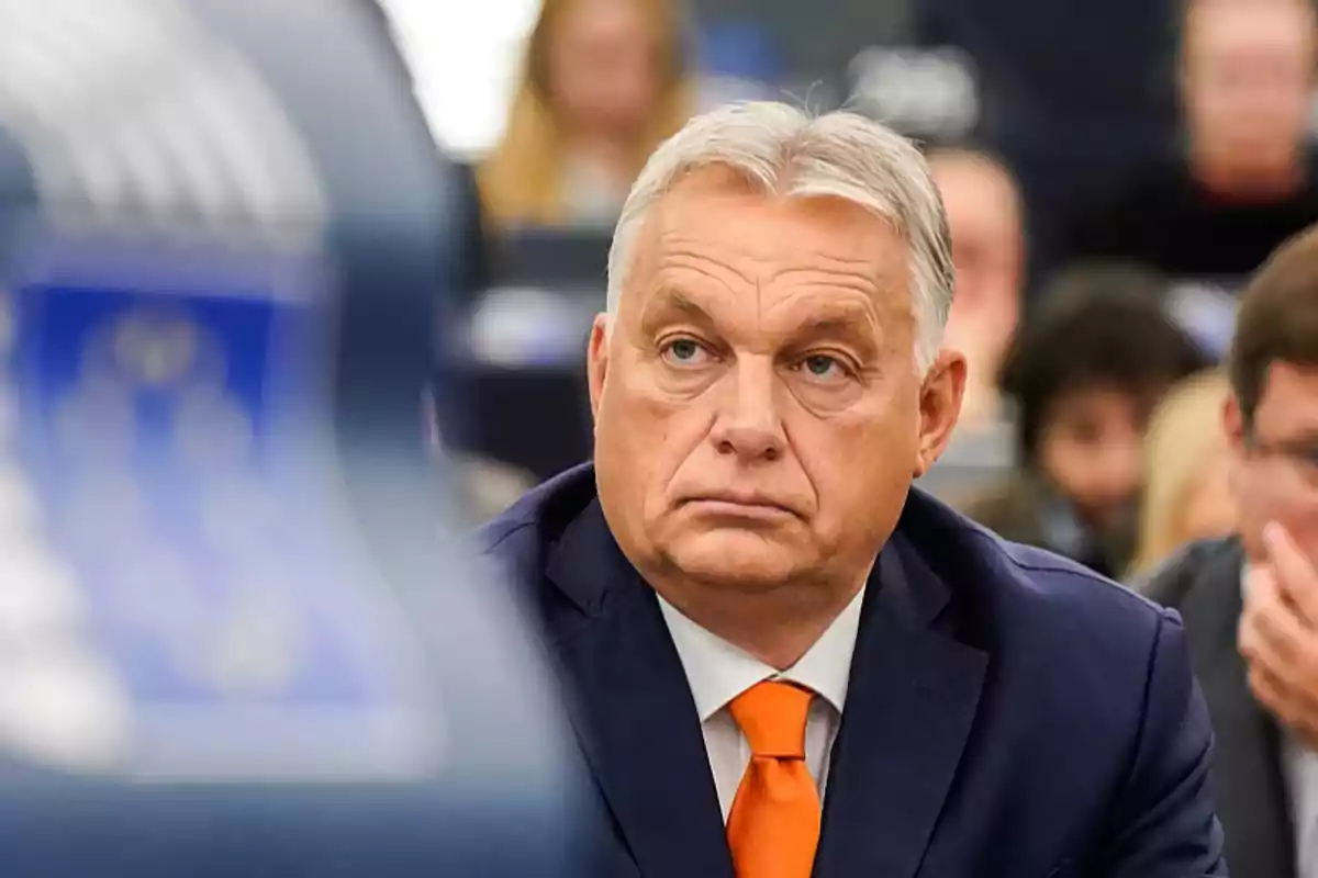 Un hombre de cabello canoso y traje oscuro con corbata naranja en un entorno de conferencia.