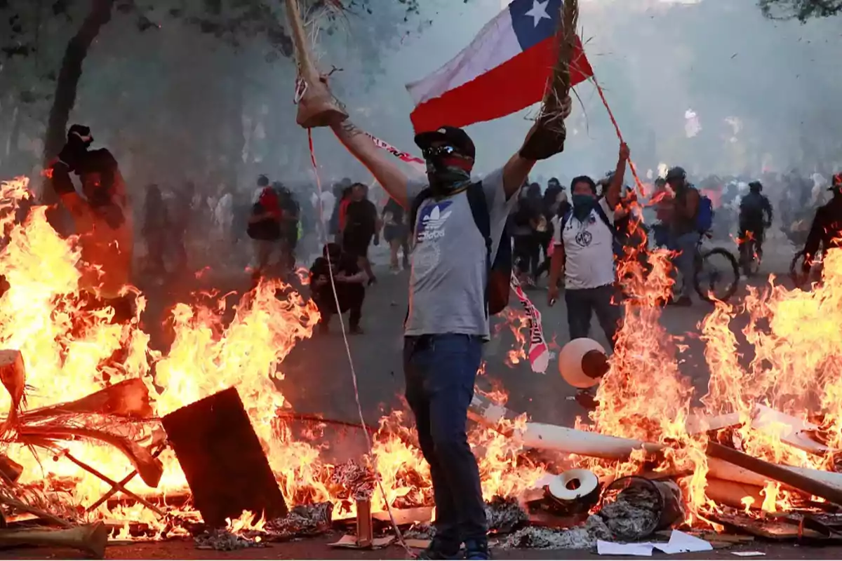 Una persona sostiene una bandera chilena frente a una barricada en llamas durante una protesta con más personas al fondo.