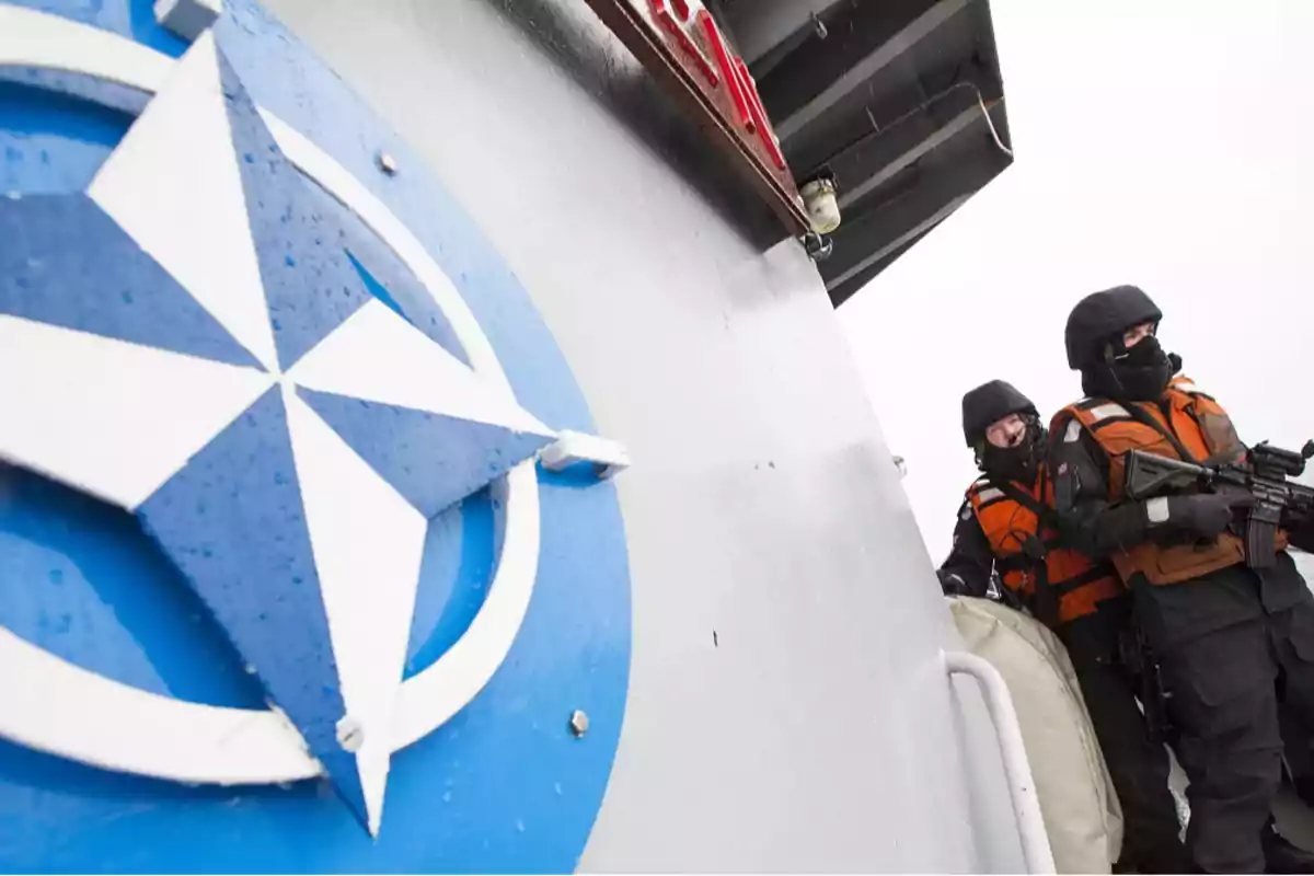 Soldados con equipo táctico junto a un barco con el emblema de la OTAN.