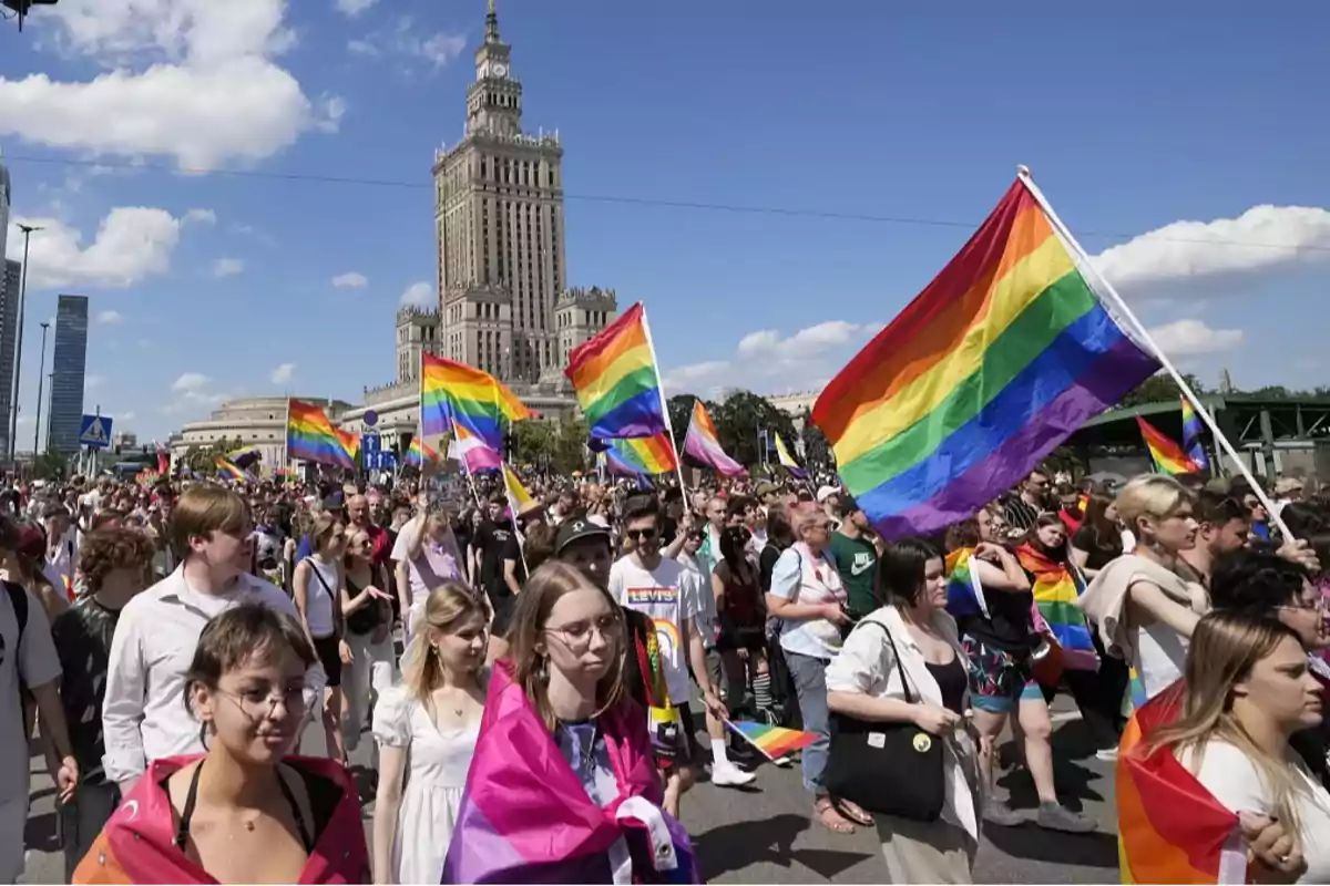 Personas marchando en un desfile del orgullo LGBTQ+ con banderas arcoíris en una ciudad con un edificio alto de fondo.