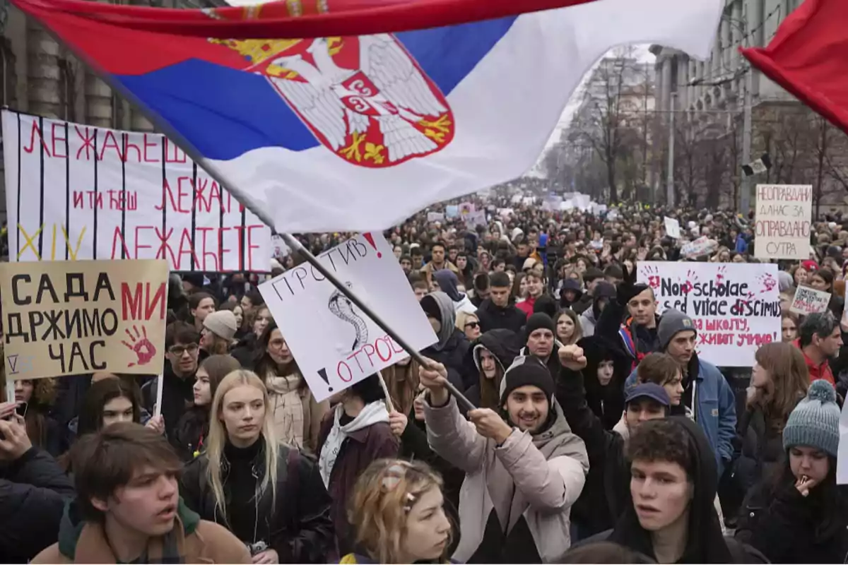 Una multitud de personas participa en una manifestación, algunas sosteniendo pancartas con mensajes escritos en serbio, mientras una gran bandera de Serbia ondea sobre ellos.