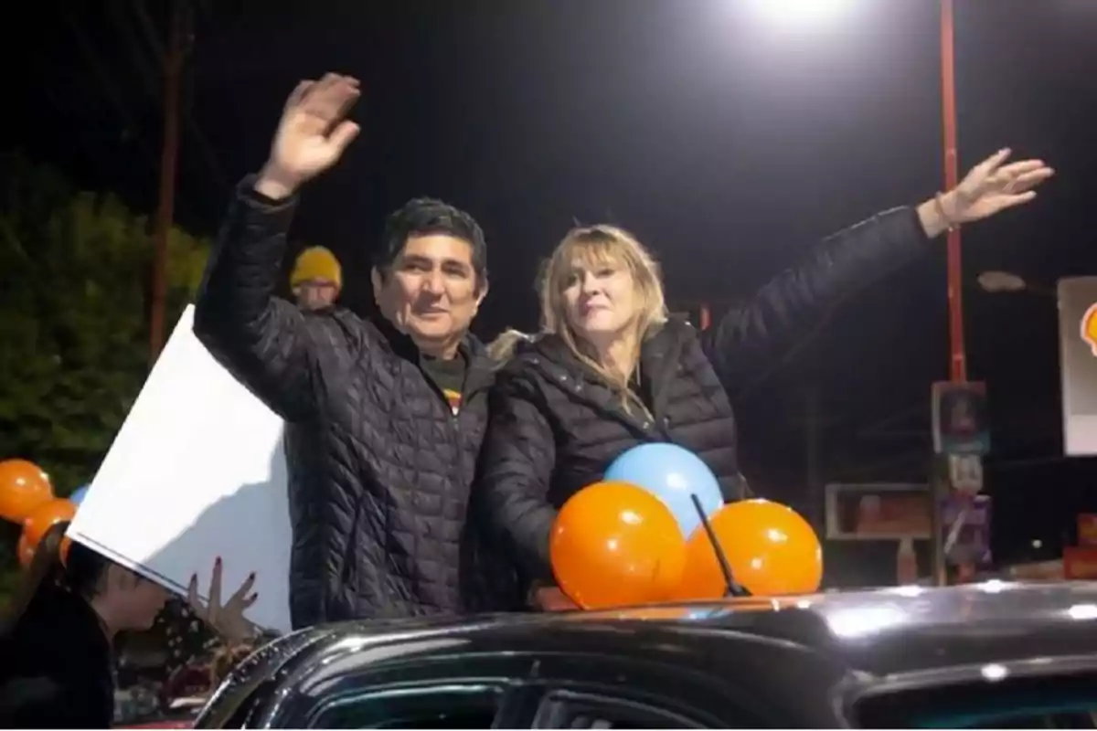 Dos personas saludan desde un vehículo decorado con globos naranjas y azules durante un evento nocturno.