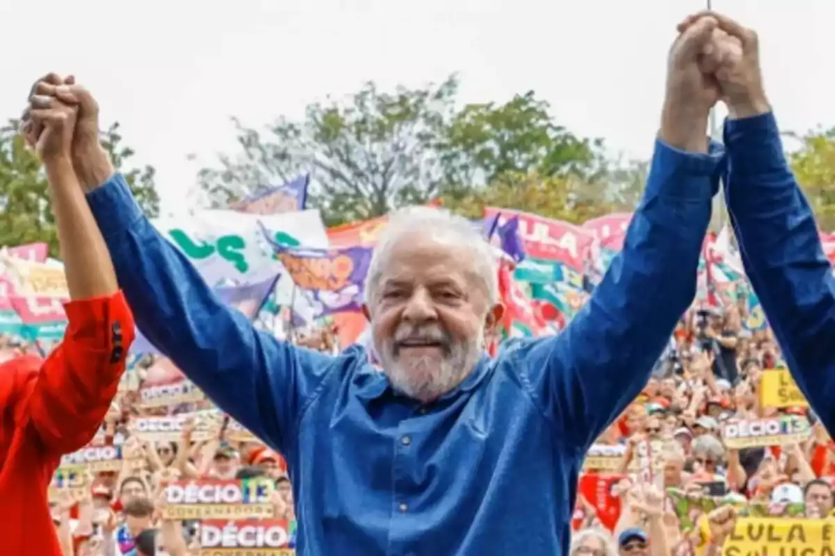 Persona mayor con barba blanca levantando los brazos en un evento público rodeado de una multitud con pancartas y banderas coloridas.