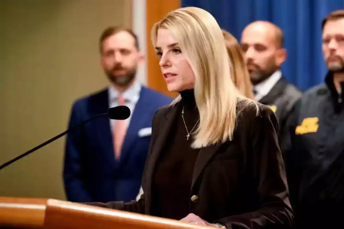 A blonde woman speaking at a podium with a microphone while several people stand in the background.