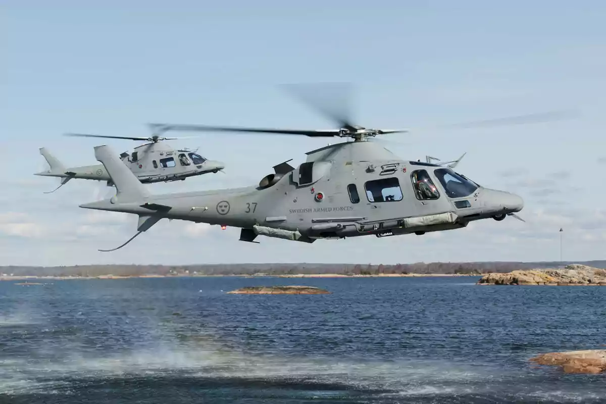 Dos helicópteros militares grises volando sobre un cuerpo de agua con un paisaje de fondo.