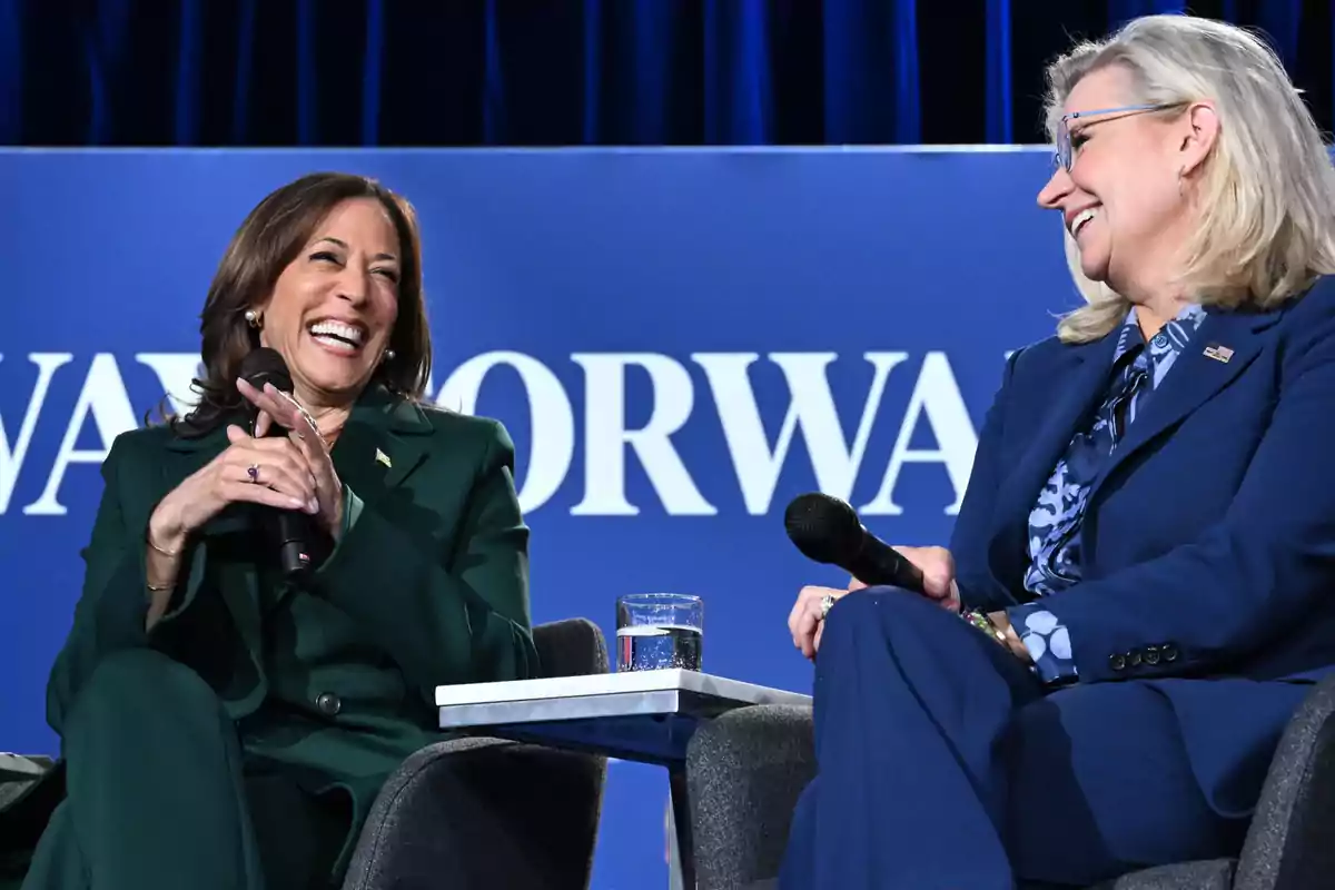 Dos mujeres sentadas en un escenario, sonriendo y sosteniendo micrófonos, con un fondo azul que tiene texto parcialmente visible.