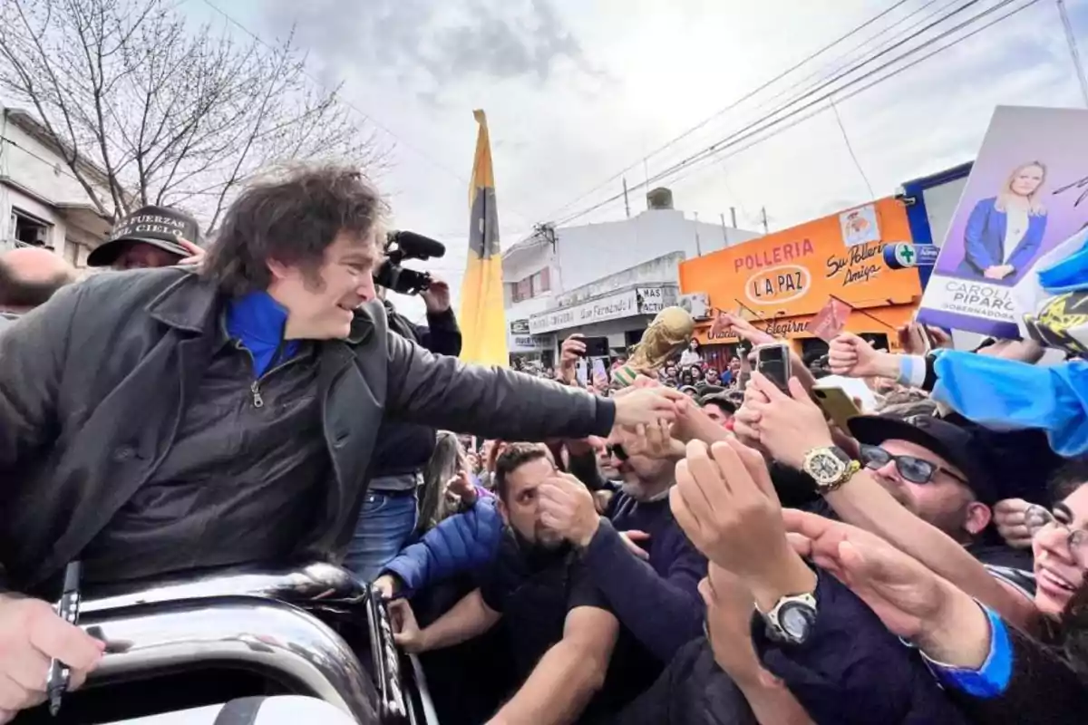 Una persona con chaqueta oscura saluda a una multitud que lo rodea mientras sostiene un trofeo dorado en un entorno urbano con carteles y banderas.