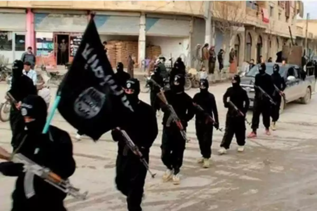 Un grupo de personas vestidas de negro marchando por una calle con armas y una bandera negra.