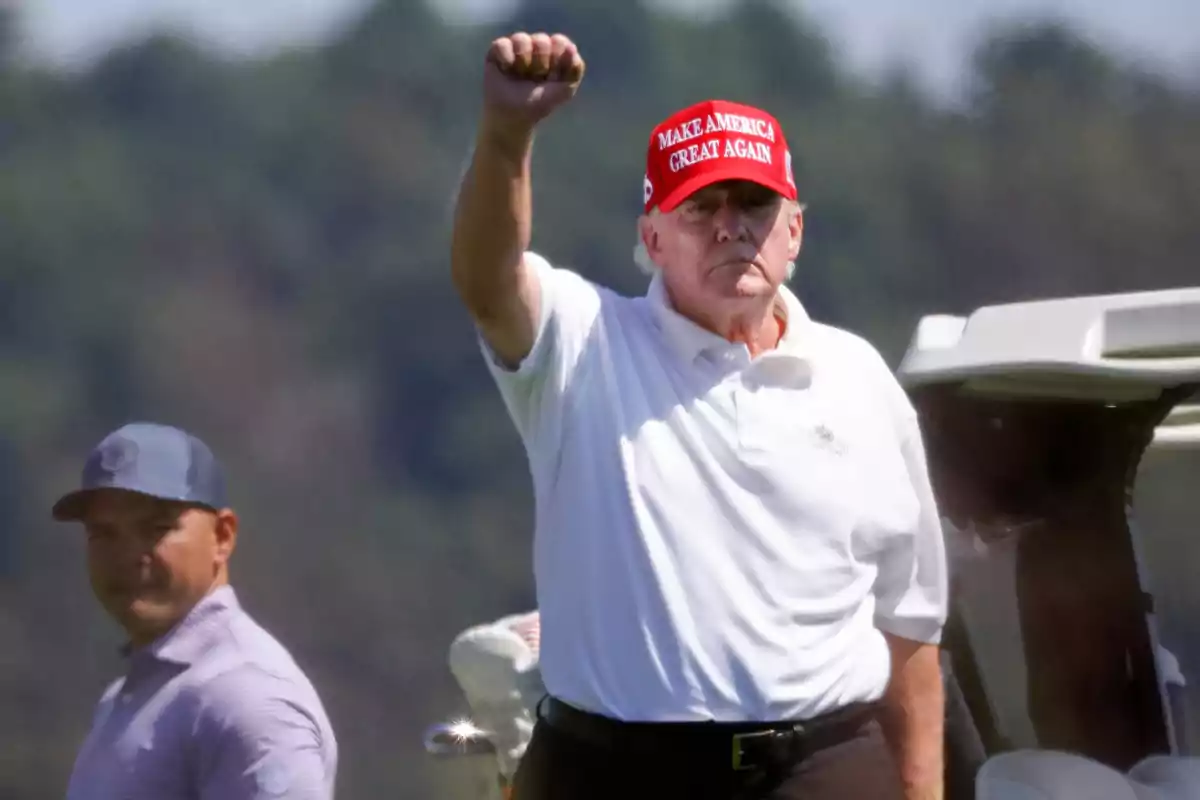 Un hombre con una gorra roja que dice "Make America Great Again" levanta el puño mientras está de pie al aire libre, con otra persona en el fondo.