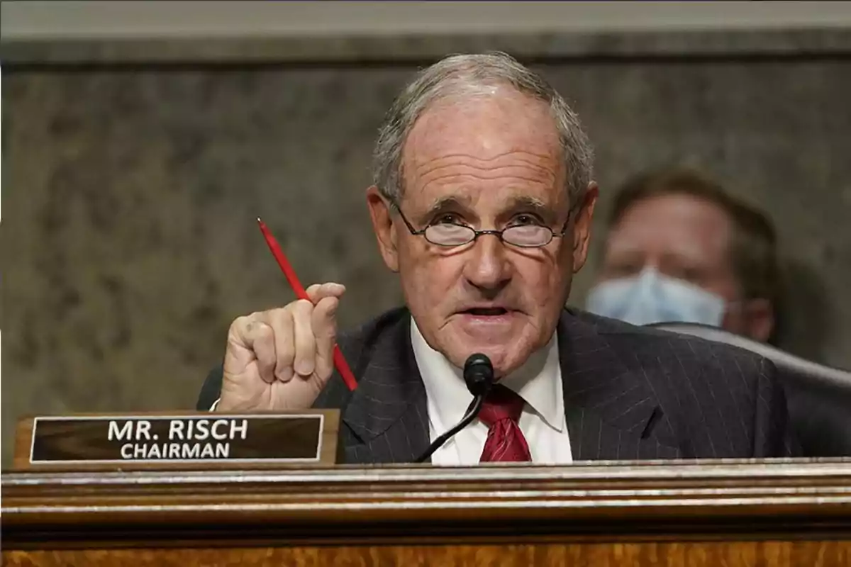 An older man with glasses holds a red pen while speaking into a microphone at a formal meeting.