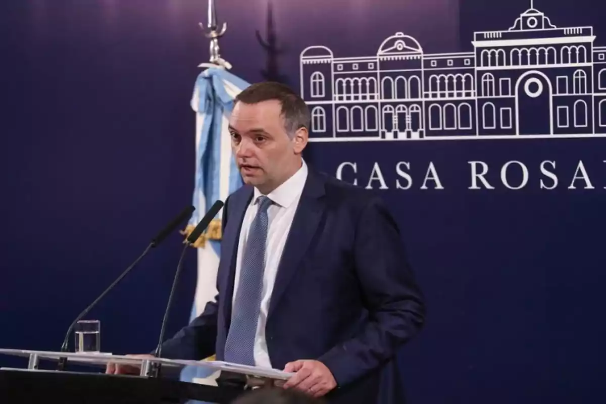 Un hombre en traje hablando en un podio con un fondo que muestra un dibujo de un edificio y las palabras Casa Rosada.